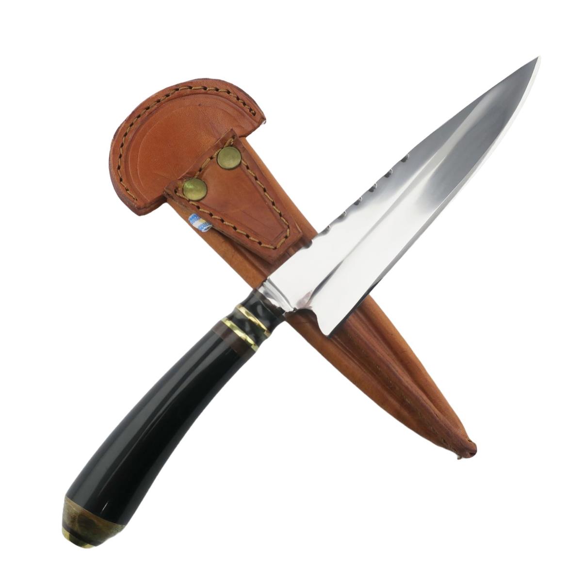 Un cuchillo de acero inoxidable Cow 6.3in Dark con mango negro y detalles de latón, con una hoja afilada y puntiaguda con dentados en el lomo, parcialmente enfundado en una funda de cuero marrón claro con remaches de latón.